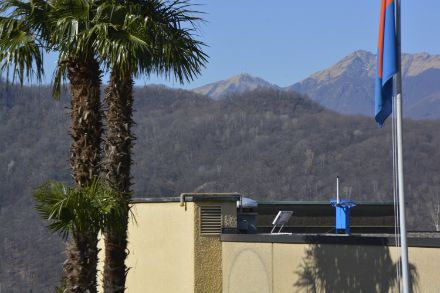Fig. 3: Il sensore blu della grandine è in armonia cromatica con la bandiera e il cielo blu del cantone meridionale della Svizzera. Ma anche in Ticino può grandinare. Il sensore grandine sul tetto della casa e il radar meteorologico sul Monte Lema sullo sfondo sono pronti 24 ore su 24 a rilevare la grandine. Fonte: MeteoSvizzera