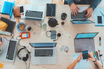 L'immagine mostra un grande tavolo di legno con diversi laptop, smartphone e materiali di lavoro, mentre le persone lavorano e interagiscono.