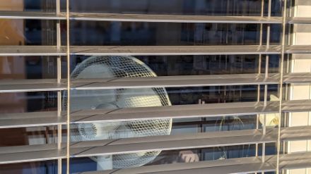  	Sguardo attraverso una finestra con le tapparelle semi-abbassate. Nella stanza è presente un ventilatore vicino alla finestra.