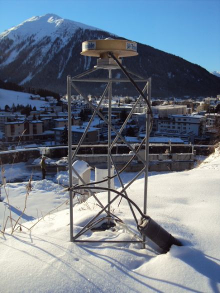 In primo piano nell’immagine lo strumento di misurazione GPS, un disco metallico montato su una struttura portante in metallo. Sullo sfondo è riconoscibile la località di Davos.