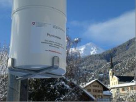 In primo piano un pluviometro, sullo sfondo i boschi e le montagne innevate.