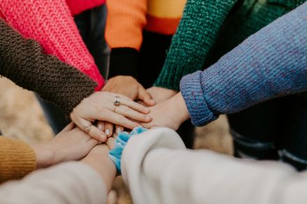 Diverse mani sono sovrapposte in segno di coesione. Le persone indossano maglioni colorati, alcune mani sono decorate con anelli. L'immagine irradia comunità e spirito di squadra.